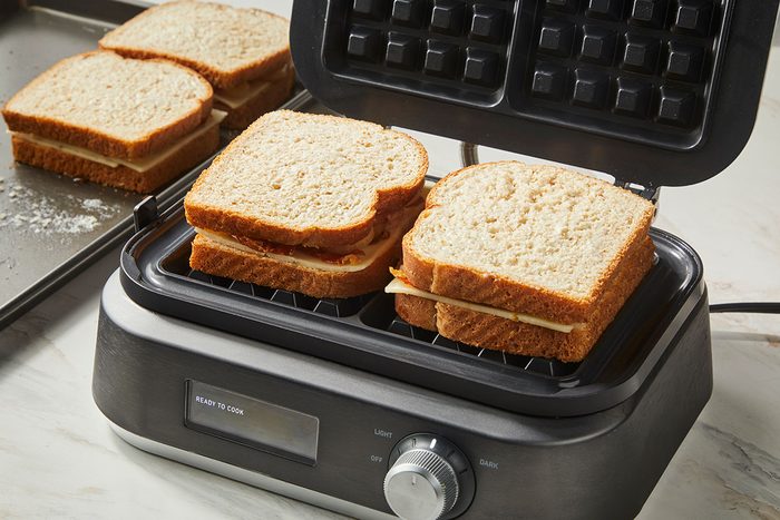 3/4th shot of wo sandwiches on wheat bread are being grilled in an open panini press, while two more sandwiches sit on a baking tray nearby; The scene is set on a light countertop