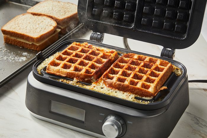 3/4th shot of An open waffle maker with two cooked square waffles inside sits on a countertop; In the background, slices of bread are placed on a baking tray; Steam rises from the waffle maker