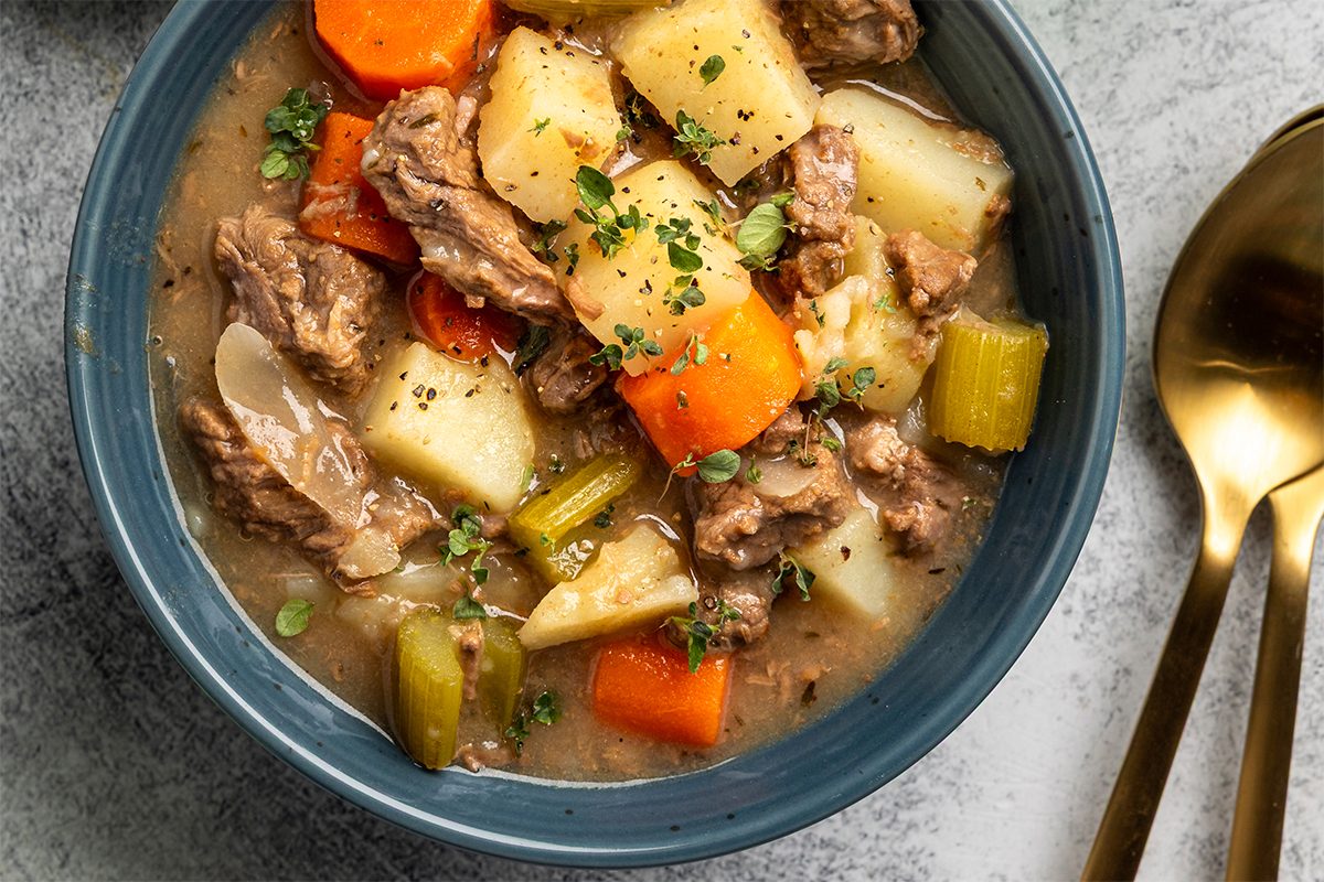 A bowl of hearty beef stew with chunks of beef, potatoes, carrots, celery, and herbs, served with a spoon and fork on the side.