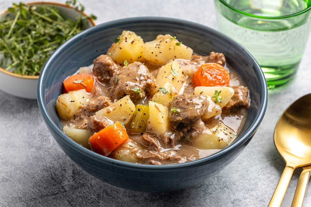 A bowl of beef stew with chunks of beef, potatoes, and carrots in a thick broth, garnished with herbs. A small bowl of greens and a green glass are nearby, with a spoon and fork on the table.