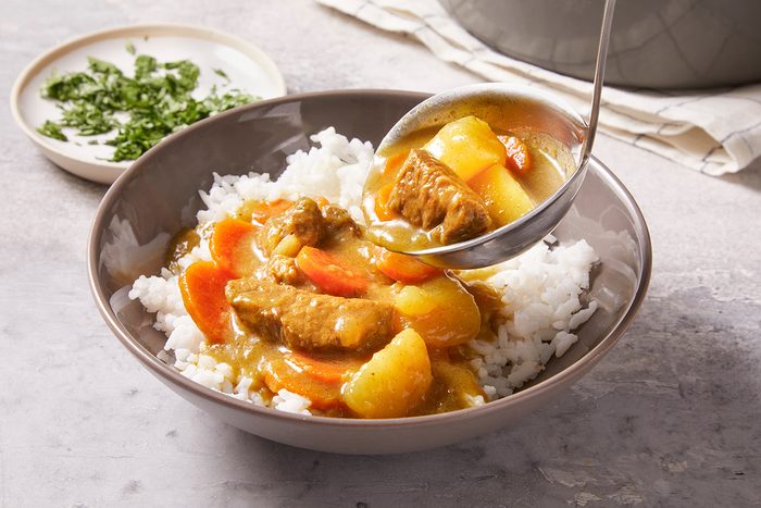 A bowl of white rice topped with Japanese curry containing beef, potatoes, and carrots, being served with a ladle. A small plate with chopped herbs is in the background.