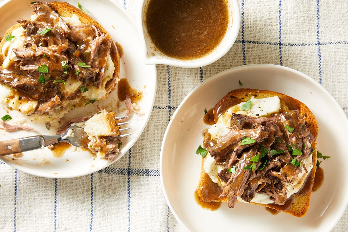 Overhead shot of two plates of Beef Manhattan served with gravy boats and utensils on a neutral tablecloth.