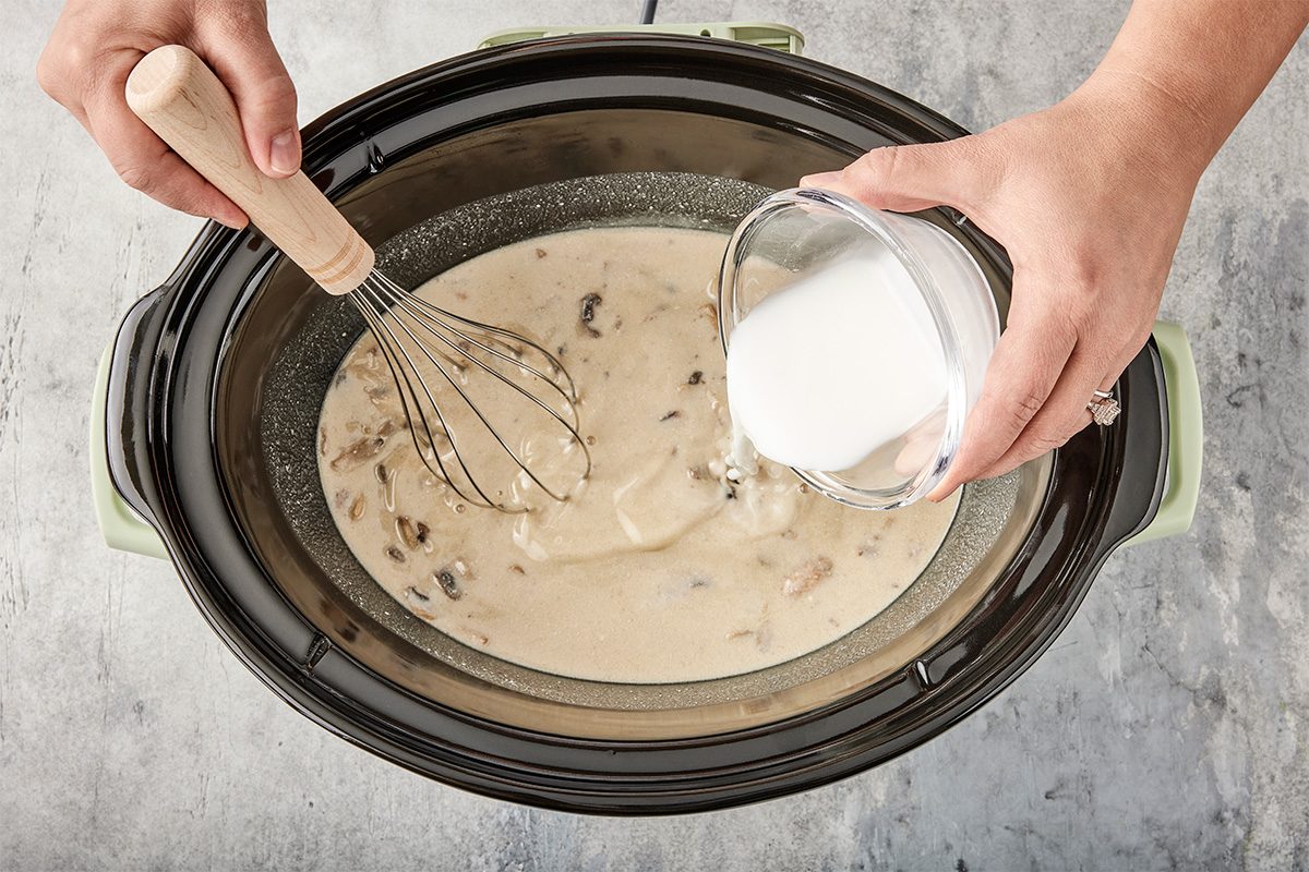 A person whisking a creamy mixture with mushrooms in a slow cooker while pouring a white liquid from a small bowl into it.