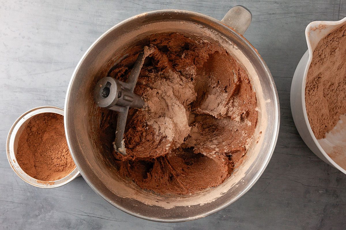 Overhead shot of mixing bowl holds chocolate batter being blended and sits on a gray countertop Containers of cocoa powder surround the bowl