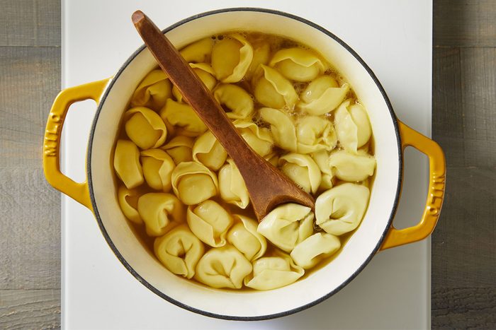 Overhead shot of cheese tortellini boiling in a white Dutch oven filled with broth. A wooden spoon rests inside, stirring the pasta as it cooks.