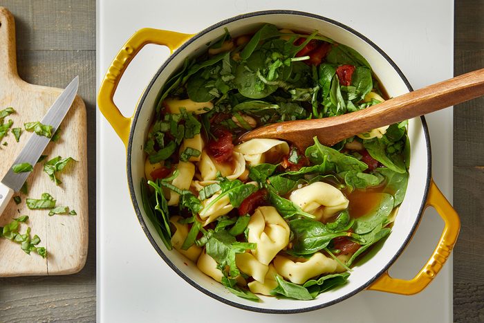 Overhead shot of fresh spinach leaves being stirred into the Dutch oven of hot soup, with a wooden spoon mixing the greens into the broth.