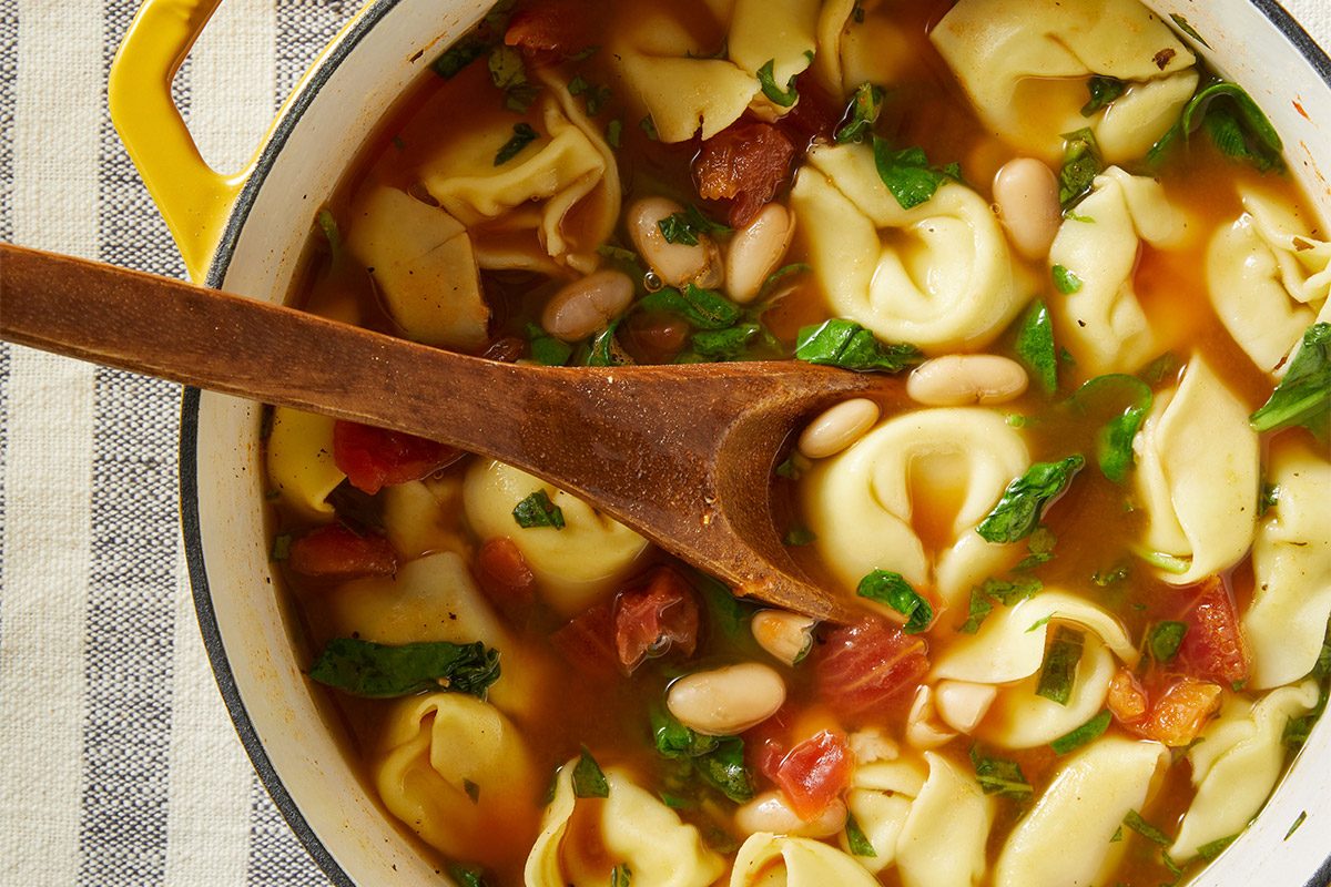 Vertical shot of Christmas Tortellini & Spinach Soup simmering in a Dutch oven, with visible tortellini, spinach, tomatoes, and beans. Asiago cheese and herbs sit nearby on the table.