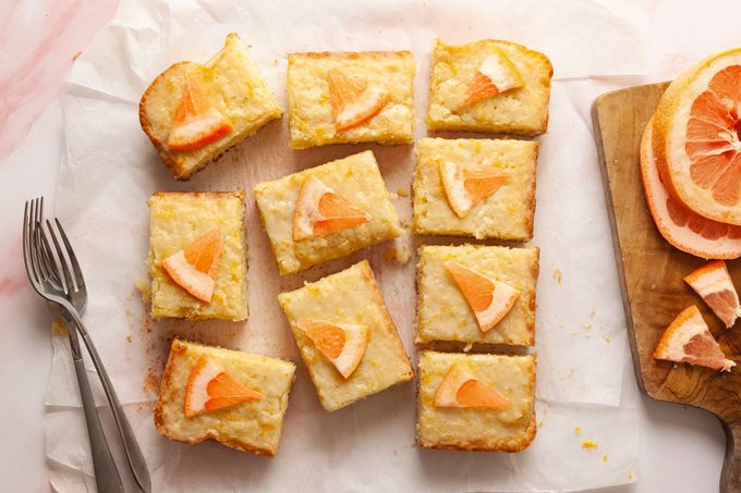 Top view shot of French Blondies—nine rectangular citrus dessert bars topped with grapefruit slices arranged on parchment paper on a pink surface; with a cutting board of grapefruit slices in the corner and two forks nearby;