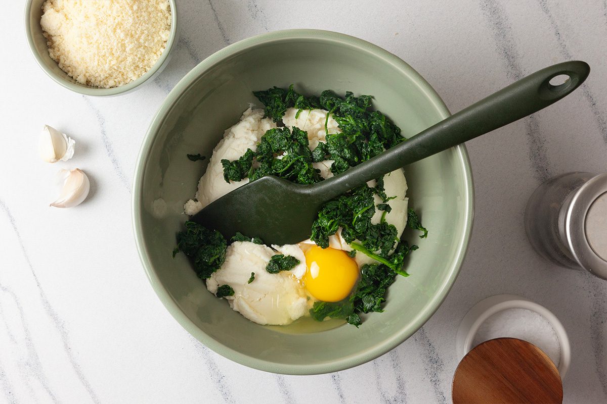 Overhead shot of a green bowl containing ricotta cheese, a raw egg, and chopped spinach being mixed with a green spatula; Nearby are bowls of grated cheese, salt, and two garlic cloves on a white countertop