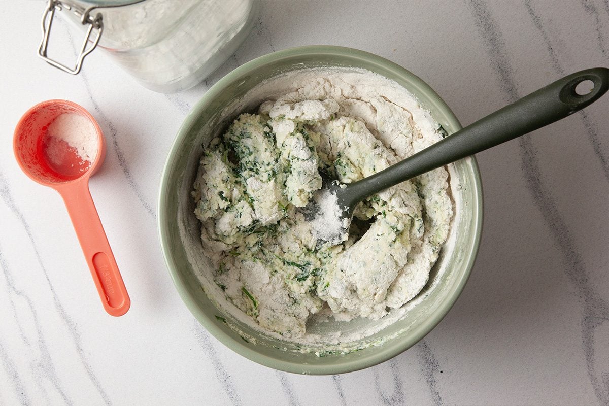 Overhead shot of a green mixing bowl with dough containing green herbs or vegetables being stirred with a black spatula, next to a red measuring cup on a marble countertop