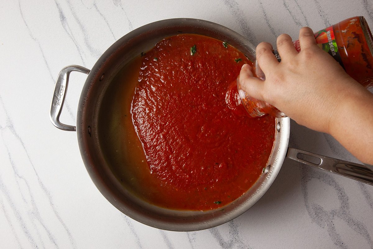 Overhead shot of a hand pouring tomato sauce from a jar into a large, shallow pan with handles on a white marble countertop