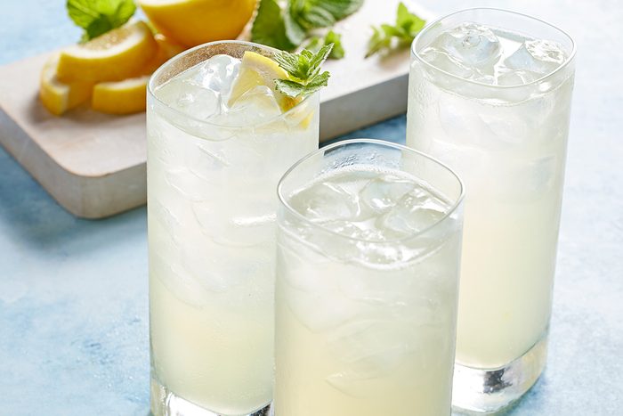 Three tall glasses of iced lemonade are placed on a light blue surface, garnished with lemon wedges and mint. In the background, sliced lemons and mint leaves rest on a wooden cutting board.