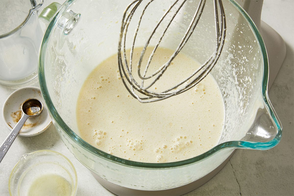 A glass mixing bowl filled with a frothy, light batter sits on a countertop next to a mixer whisk attachment. Nearby are a measuring spoon, a small bowl with liquid, and a glass cup with more liquid.