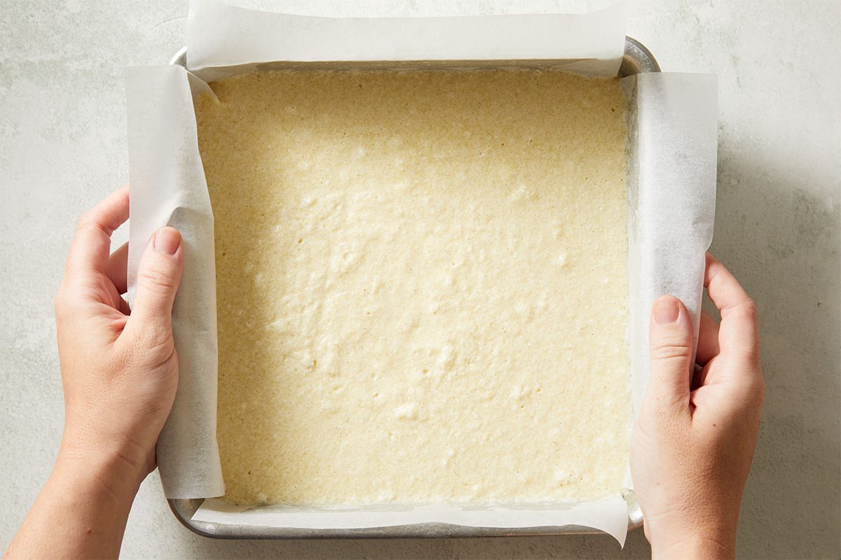 Two hands hold a square baking pan lined with parchment paper and filled with an even layer of pale, uncooked batter, ready to be baked.