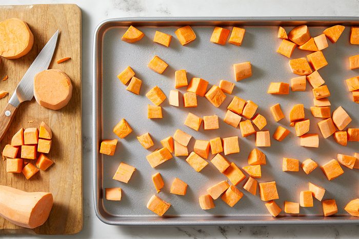 Overhead shot of cubed sweet potatoes spread on a baking sheet with a wooden cutting board and knife beside them.