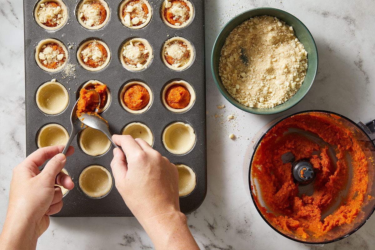 Overhead shot of hands spooning sweet potato mixture into mini tart shells with a bowl of topping beside the tray.