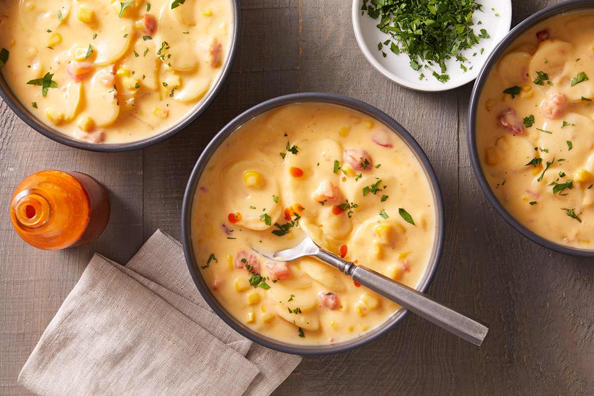 Three bowls of creamy soup with visible corn, potatoes, and herbs, garnished with chopped parsley. A small bowl of extra herbs and an orange bottle are nearby on a gray surface, next to a folded beige napkin.