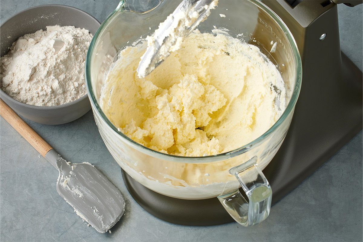 High-angle shot of a stand mixer bowl containing creamed butter and sugar with the mixing paddle attached, alongside a bowl of flour and a gray spatula resting on the countertop;