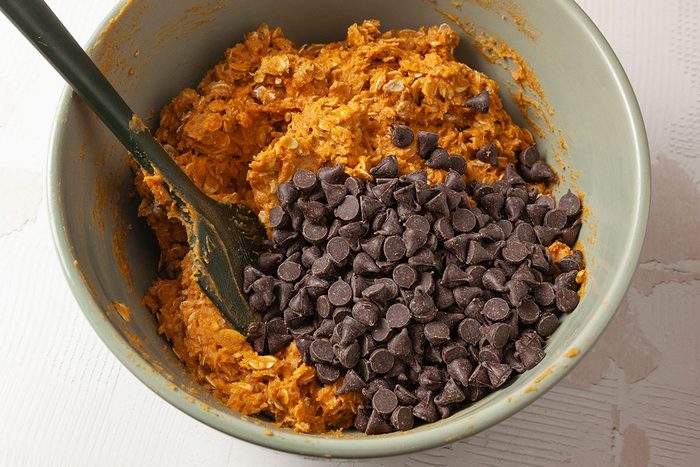 Closeup shot of a mixing bowl filled with cookie dough mixed with oats and topped with a large pile of chocolate chips; a spoon is partially submerged in the mixture.