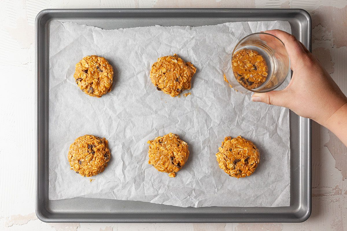 Overhead shot of a hand pressing a cookie dough ball flat with a glass on a parchment-lined baking tray; five unbaked cookies are evenly spaced and ready to bake.