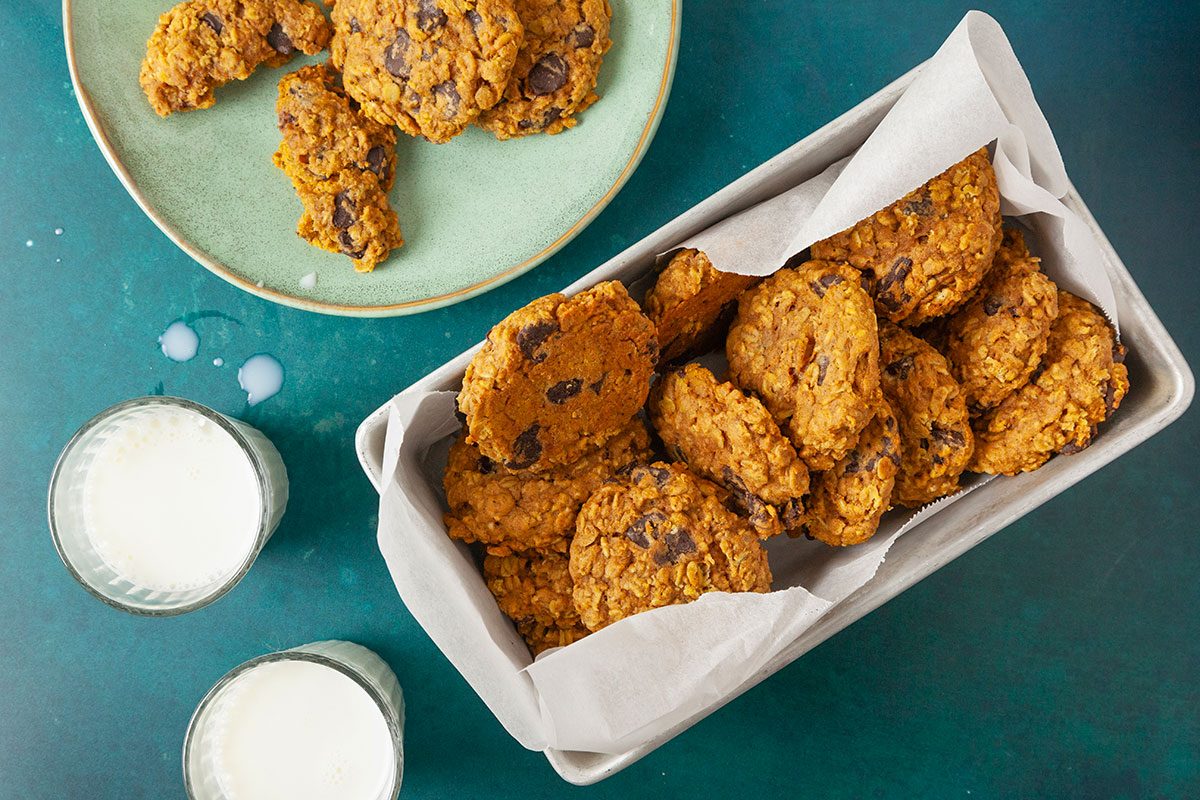 Overhead shot of a rectangular dish lined with parchment paper holding Pumpkin Oatmeal Cookies; two glasses of milk and more cookies on a plate sit nearby on a teal surface.