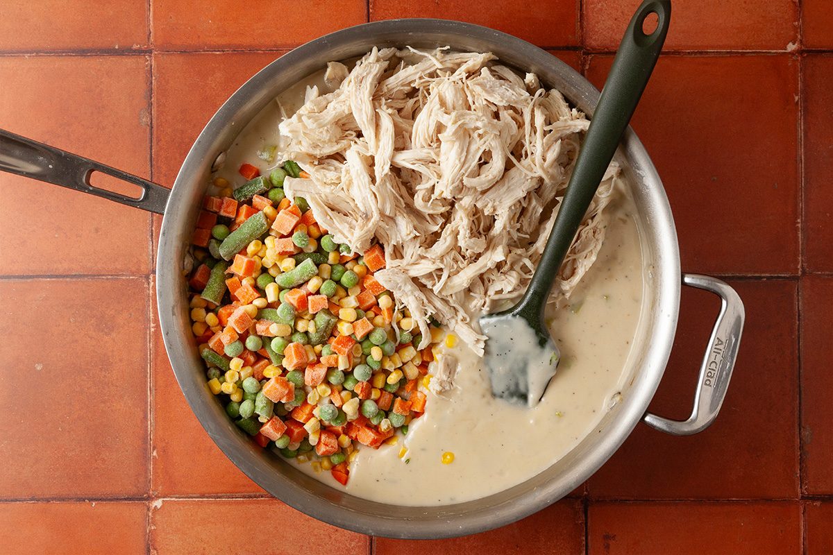 Overhead shot of a skillet on a tiled surface containing shredded chicken, mixed frozen vegetables, and creamy sauce, with a spatula resting inside