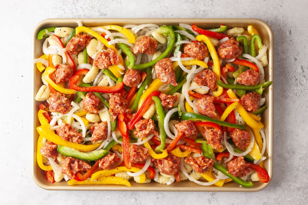 Overhead shot of uncooked sausage, bell peppers, onions, and potatoes mixed and seasoned on a sheet pan on a textured background