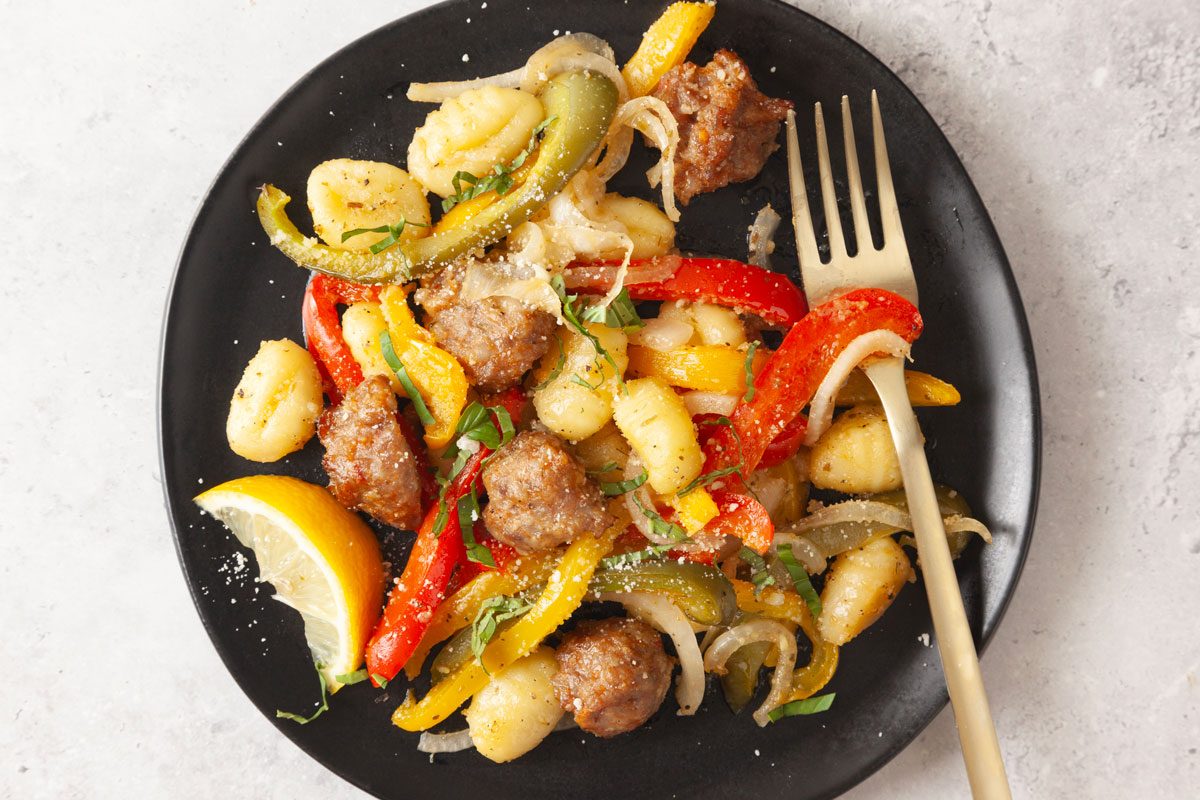 Overhead shot of Sheet-Pan Gnocchi with Sausage and Peppers served on a black plate with a single serving, garnished with lemon wedge and served with a gold fork.