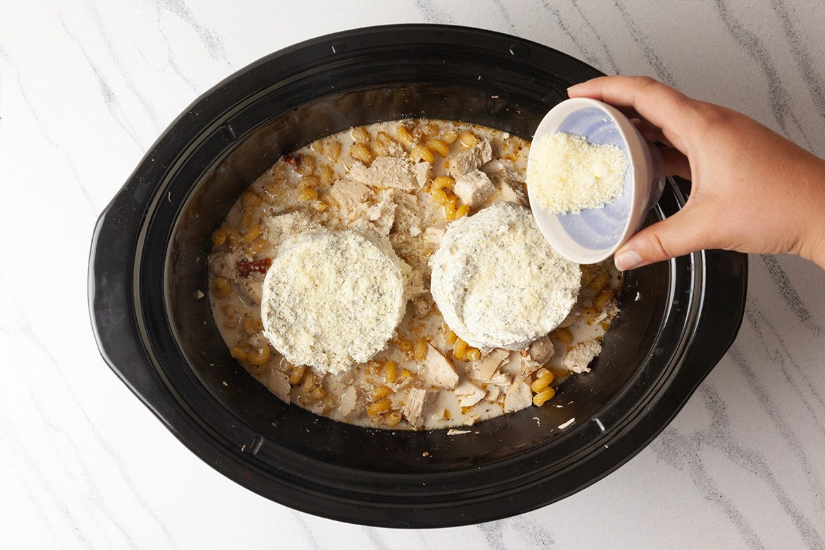 Overhead shot of a hand sprinkling shredded cheese from a small bowl into a slow cooker filled with corn, chicken, sauce, and two round mounds topped with cheese, on a white marble surface