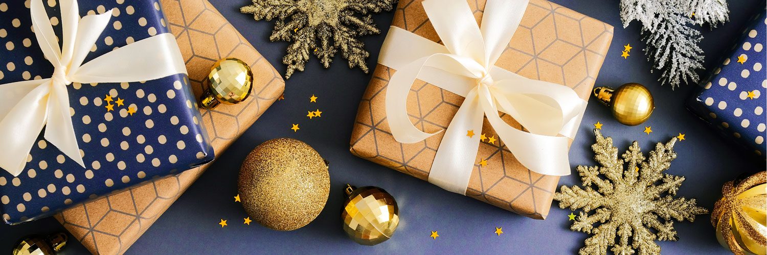 Festive flat lay with gift boxes wrapped in blue and gold with bows and ornaments and confetti on dark blue background.