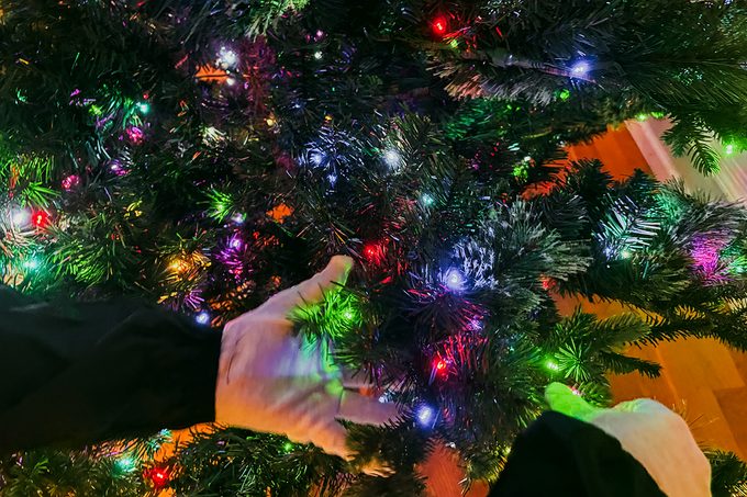 Close-up of two hands adjusting or decorating a Christmas tree with colorful, glowing lights. The tree branches are dense, and the lights are red, green, blue, and white. The scene is festive and indoors.