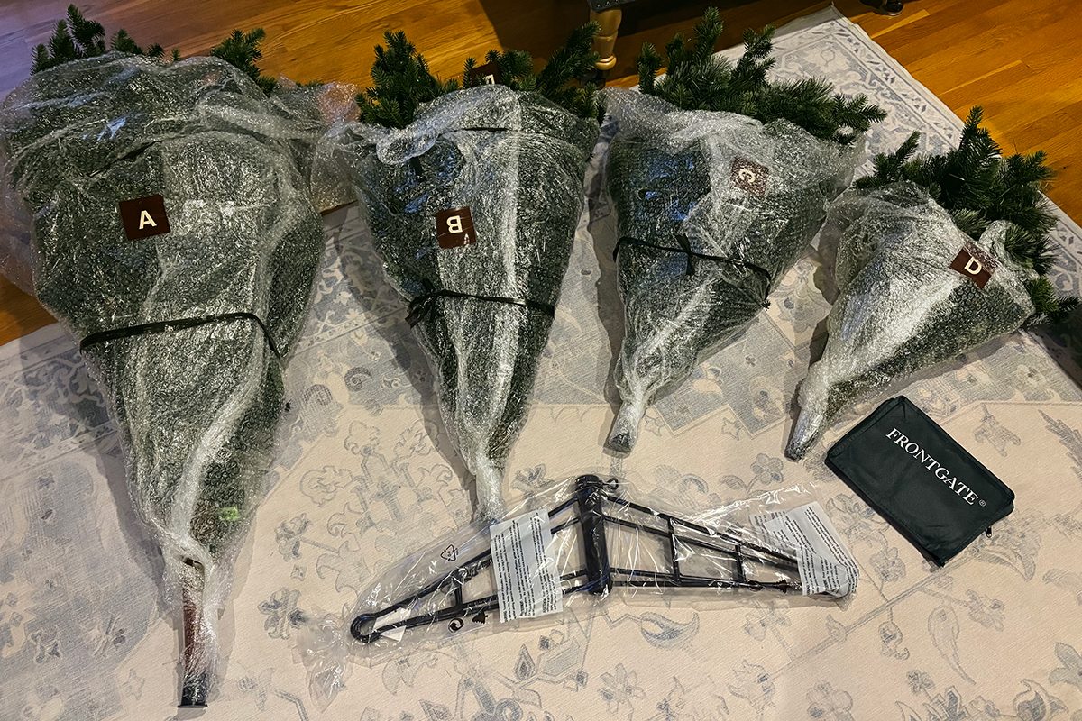 Four artificial Christmas tree sections labeled A–D are wrapped in plastic and laid out on a rug, along with a black metal tree stand and a black storage bag.