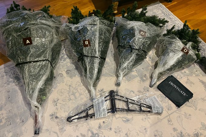 Four artificial Christmas tree sections labeled A–D are wrapped in plastic and laid out on a rug, along with a black metal tree stand and a black storage bag.