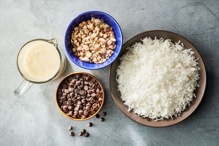 Overhead shot of a bowl of shredded coconut, a bowl of chopped nuts, a bowl of chocolate chips, and a cup of sweetened condensed milk arranged on a gray surface;