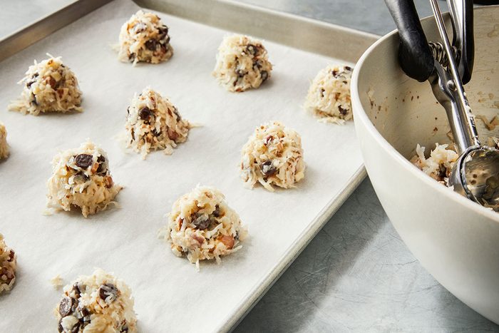 3/4 angle view of scoops of unbaked cookie dough with chocolate chips arranged on a parchment-lined baking sheet, with a mixing bowl and metal cookie scoop nearby;