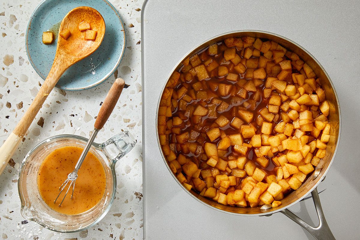 Overhead shot of a saucepan filled with diced apples in caramel sauce on a countertop, with a wooden spoon holding apple pieces, a blue plate, and a glass measuring cup of sauce with a whisk nearby;
