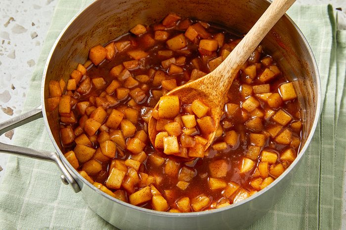 Overhead shot of a saucepan filled with diced apples cooked in a thick, brown spiced sauce, with a wooden spoon stirring the mixture as the pan rests on a light green cloth;