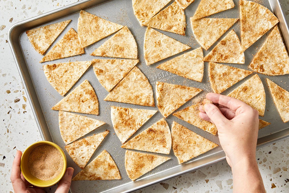 Overhead shot of a person sprinkling cinnamon-sugar over pita chips arranged on a baking sheet, holding a small bowl of the cinnamon-sugar mixture in one hand as they prepare the chips for baking;