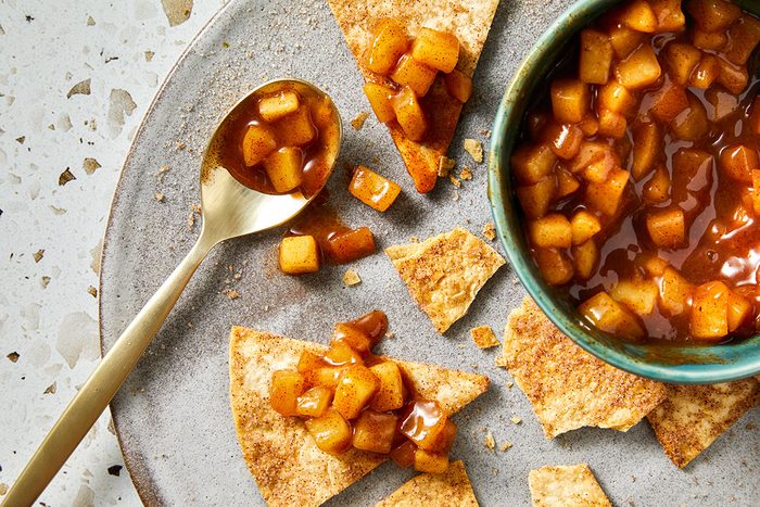 Overhead shot of apple pie dip spilled onto cinnamon-sugar pita crisps on a plate, with a gold spoon holding more of the spiced apple mixture beside the partially visible bowl;