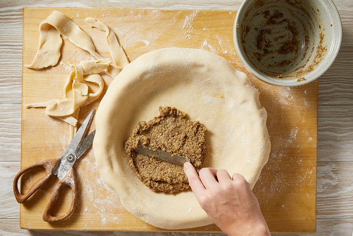 A hand spreads a brown filling into a pie crust in a round dish on a wooden board. Strips of dough, a pair of scissors, and a mixing bowl with leftover filling are nearby. Flour is scattered on the board.