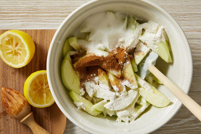 A mixing bowl filled with sliced green apples, sugar, flour, and cinnamon, next to a cutting board with halved lemons and a wooden citrus reamer.