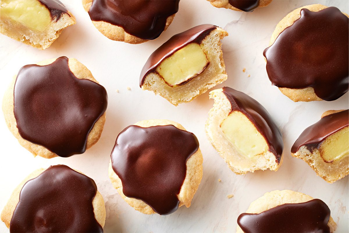 Overhead shot of Boston Cream Pie Cookies arranged on a light surface, featuring golden cookie cups with glossy chocolate tops and a few sliced open to reveal the creamy custard filling;