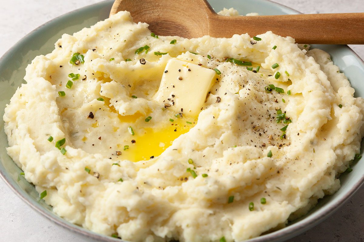 Closeup shot of Boursin Mashed Potatoes in a shallow bowl, showing their creamy texture; A pat of butter melts into the center, surrounded by cracked black pepper and sprinkled chives; A wooden spoon rests along the edge of the bowl;
