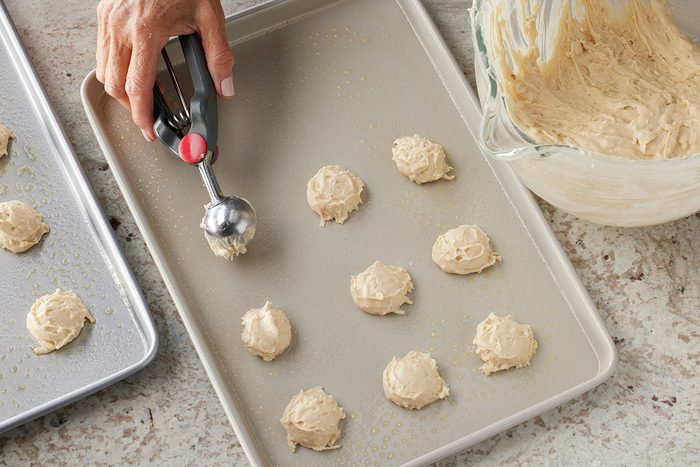 Overhead shot of a hand using a cookie scoop to place balls of cookie dough onto a greased baking sheet, with a bowl of dough and another baking sheet nearby on a countertop;