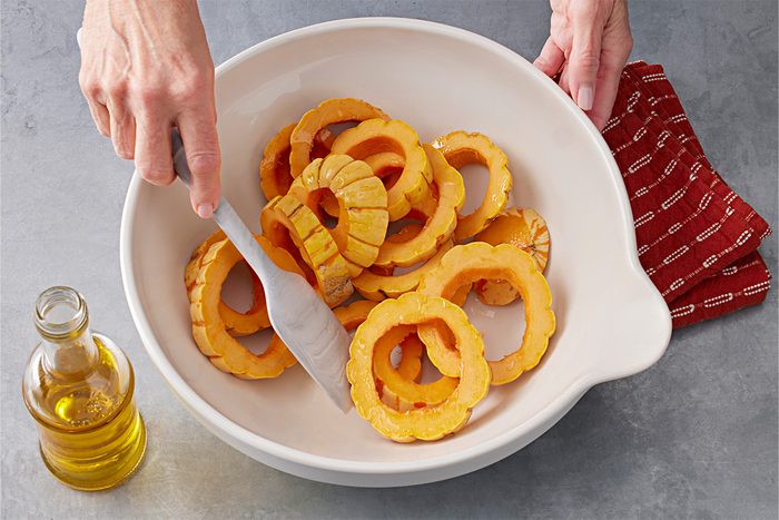 Overhead shot of a person tossing sliced delicata squash with tongs in a white bowl, with a small bottle of olive oil and a red cloth on a gray countertop nearby;