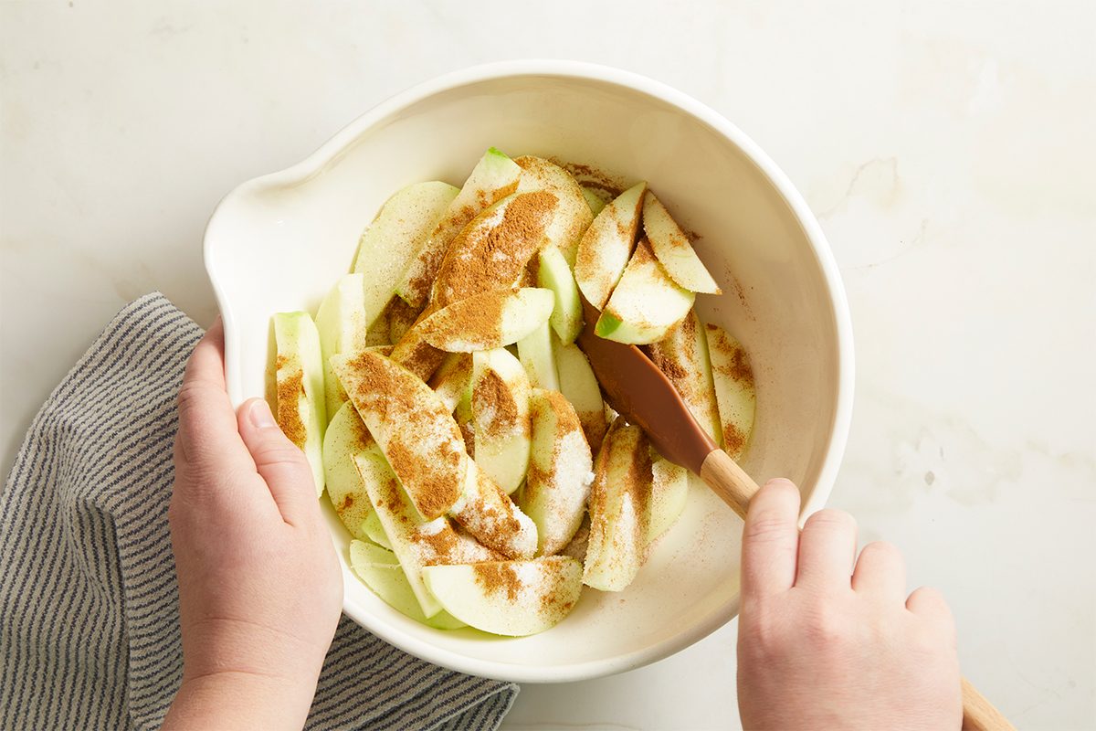 Hands stirring sliced apples with cinnamon and sugar in a large white mixing bowl with a spatula. The bowl sits on a marble surface next to a striped towel.