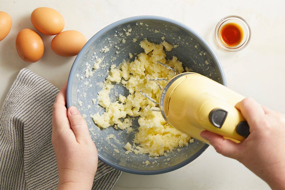 A person uses a yellow hand mixer to blend ingredients in a blue bowl. Three eggs, a small jar of vanilla extract, and a striped towel are nearby on a light counter.
