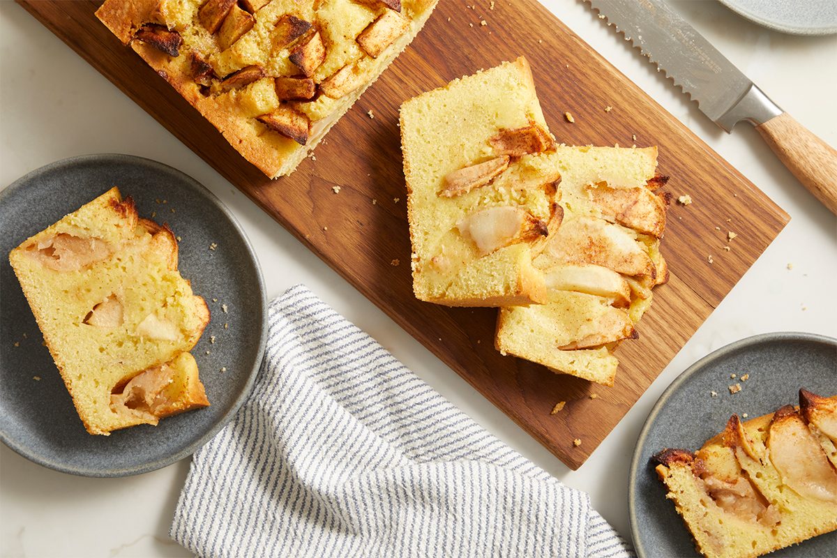 Sliced apple cake on a wooden board with two pieces on gray plates nearby, a serrated knife, and a striped kitchen towel on a white marble surface.