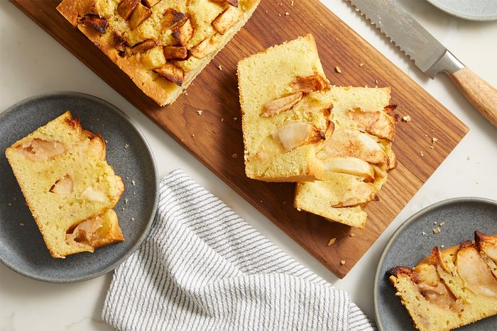 Sliced apple cake on a wooden board with two pieces on gray plates nearby, a serrated knife, and a striped kitchen towel on a white marble surface.