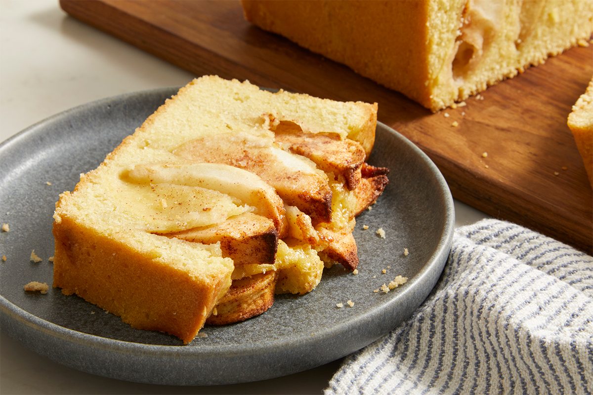 A slice of apple cake with visible apple slices and cinnamon is served on a gray plate, with more cake and a striped napkin in the background.